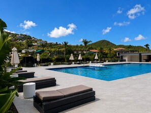 Ein Swimmingpool mit Liegestühlen und Sonnenschirmen, umgeben von Grünflächen und Gebäuden im Hintergrund.