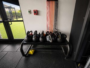 Ein Fitnessraum mit einer Bank, auf der Kettlebells liegen, einem Rauchmelder und Blick auf eine grüne Außenanlage.
