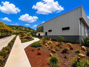 Um edifício moderno com fachada metálica, cercado por um jardim arborizado com diversas plantas e um caminho pavimentado.