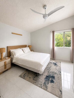 Ein Schlafzimmer mit Bett, Nachttischen, Deckenventilator, einem Fenster mit Vorhängen und Blick ins Grüne.