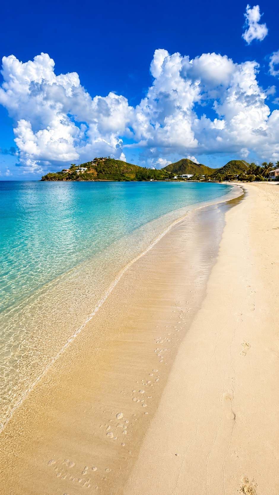 A beach with clear turquoise water, a sandy shore, and a small hill with buildings in the background.