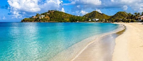 A beach with clear turquoise water, a sandy shore, and a small hill with buildings in the background.
