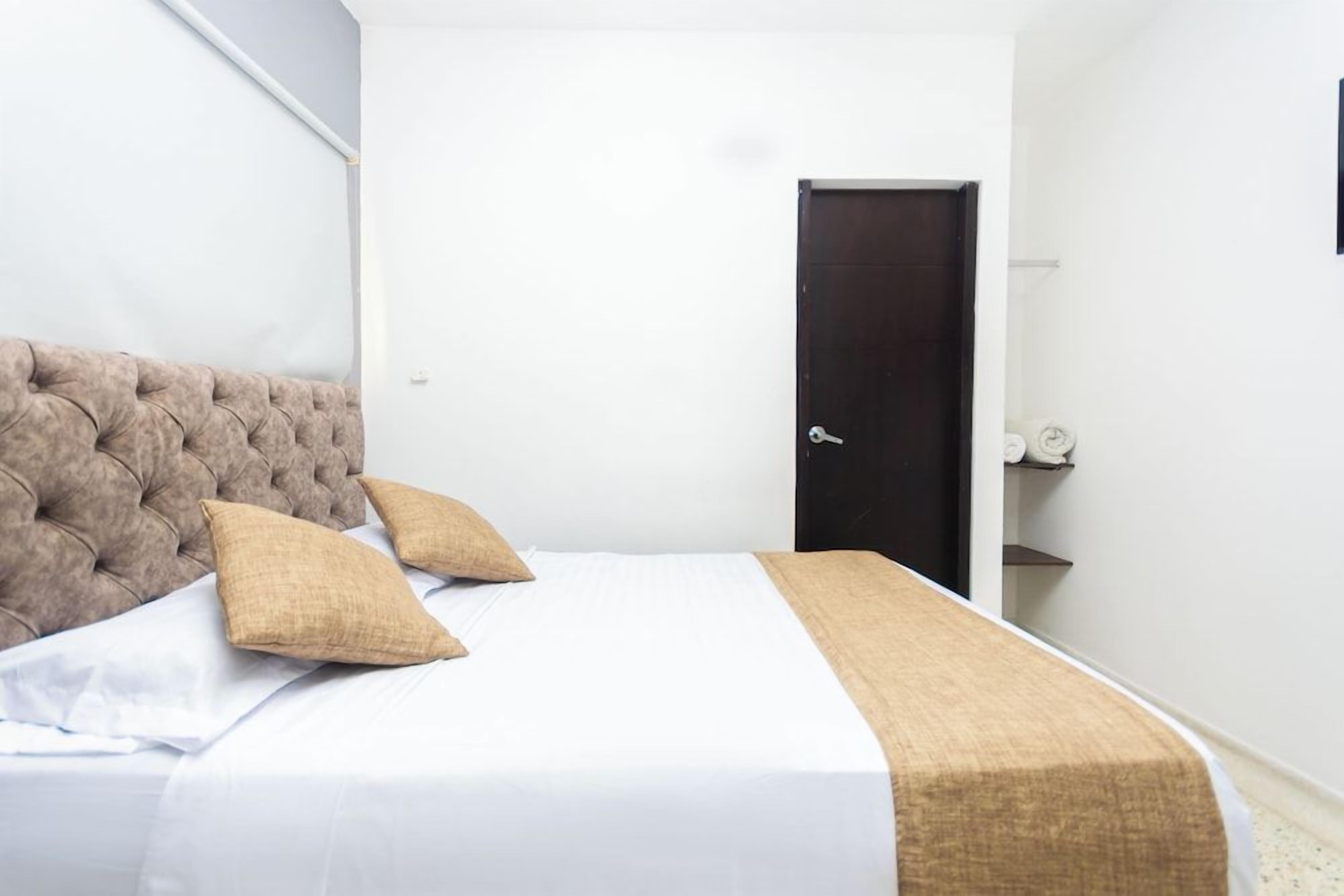 Ein Schlafzimmer mit einer gesteppten Kopfteile, einem Bett mit weißer Bettwäsche und zwei beigen Kissen, einer dunklen Tür und einem kleinen Regal mit Handtüchern.