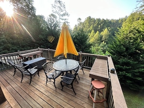 Een houten terras met een ronde tafel en stoelen, een gele parasol en een klein krukje.