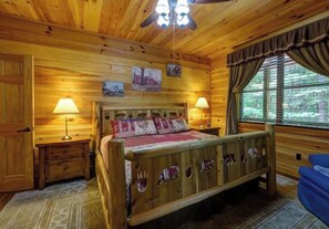 Un dormitorio de cabaña de madera con una cama, dos mesitas de noche, un ventilador de techo y una ventana con cortinas.
