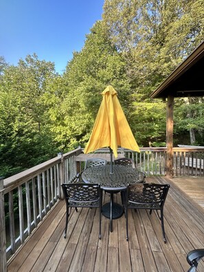 Een houten terras met een metalen tafel en stoelen, een parasol en uitzicht op bomen.