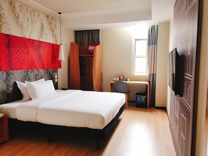 Une chambre d’hôtel comprenant un lit, un bureau avec une chaise, une armoire, un téléviseur et une fenêtre avec des rideaux.