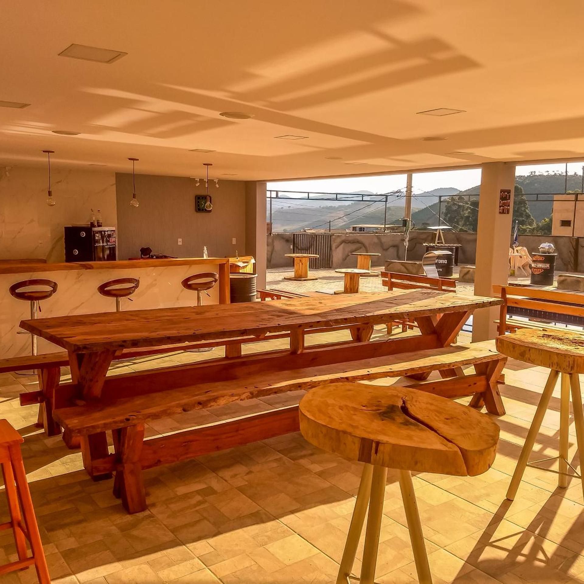 Uma área de refeições ao ar livre espaçosa, com mesas e bancos de madeira, um bar com banquetas e vista para a paisagem circundante.