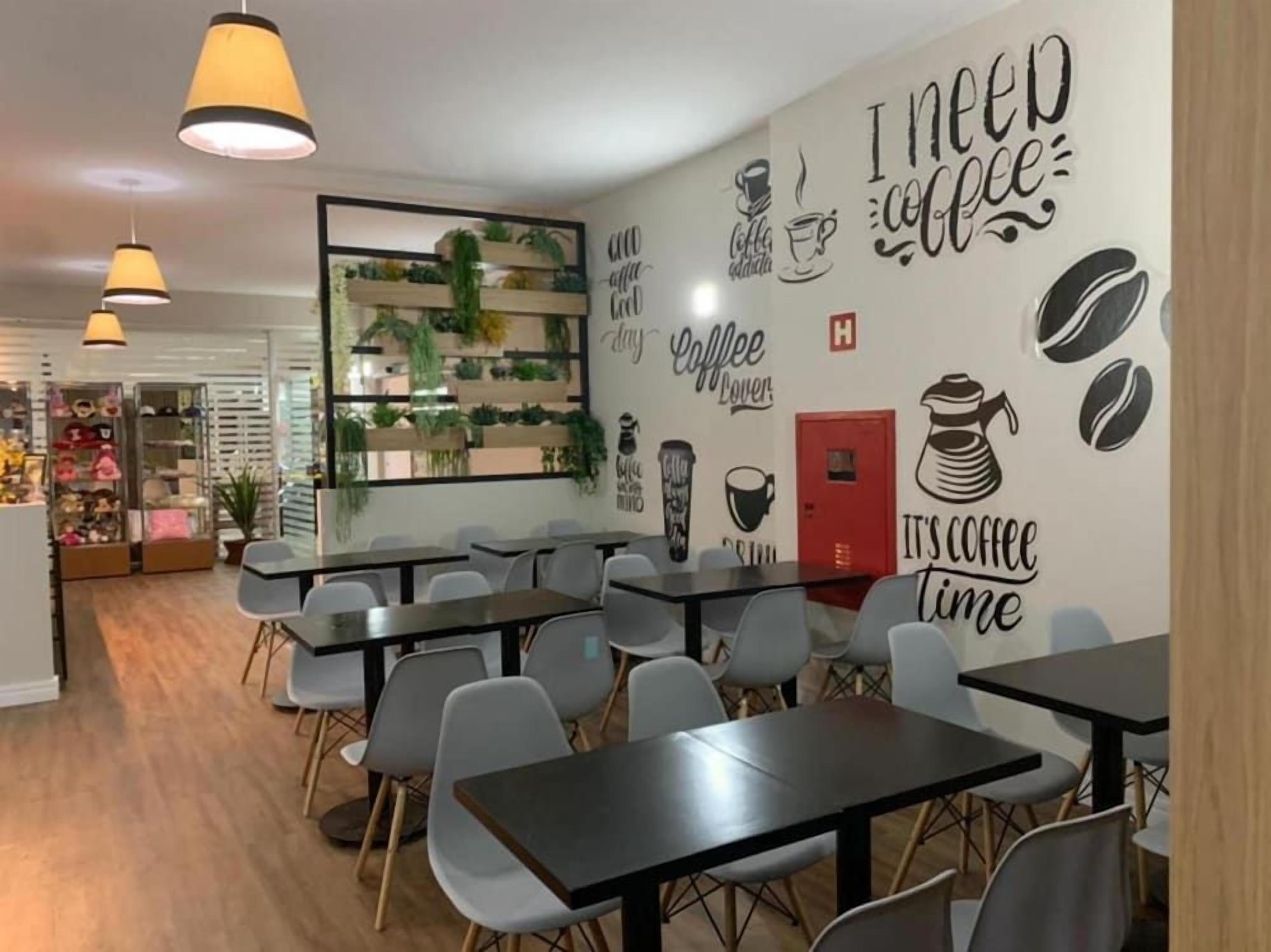 Interior de café com mesas, cadeiras e decorações nas paredes, incluindo frases e imagens relacionadas a café.