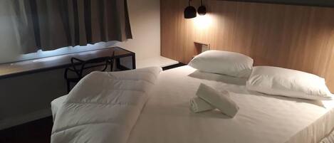 A hotel room with a bed, a desk, a chair, a lamp, and a window with curtains.