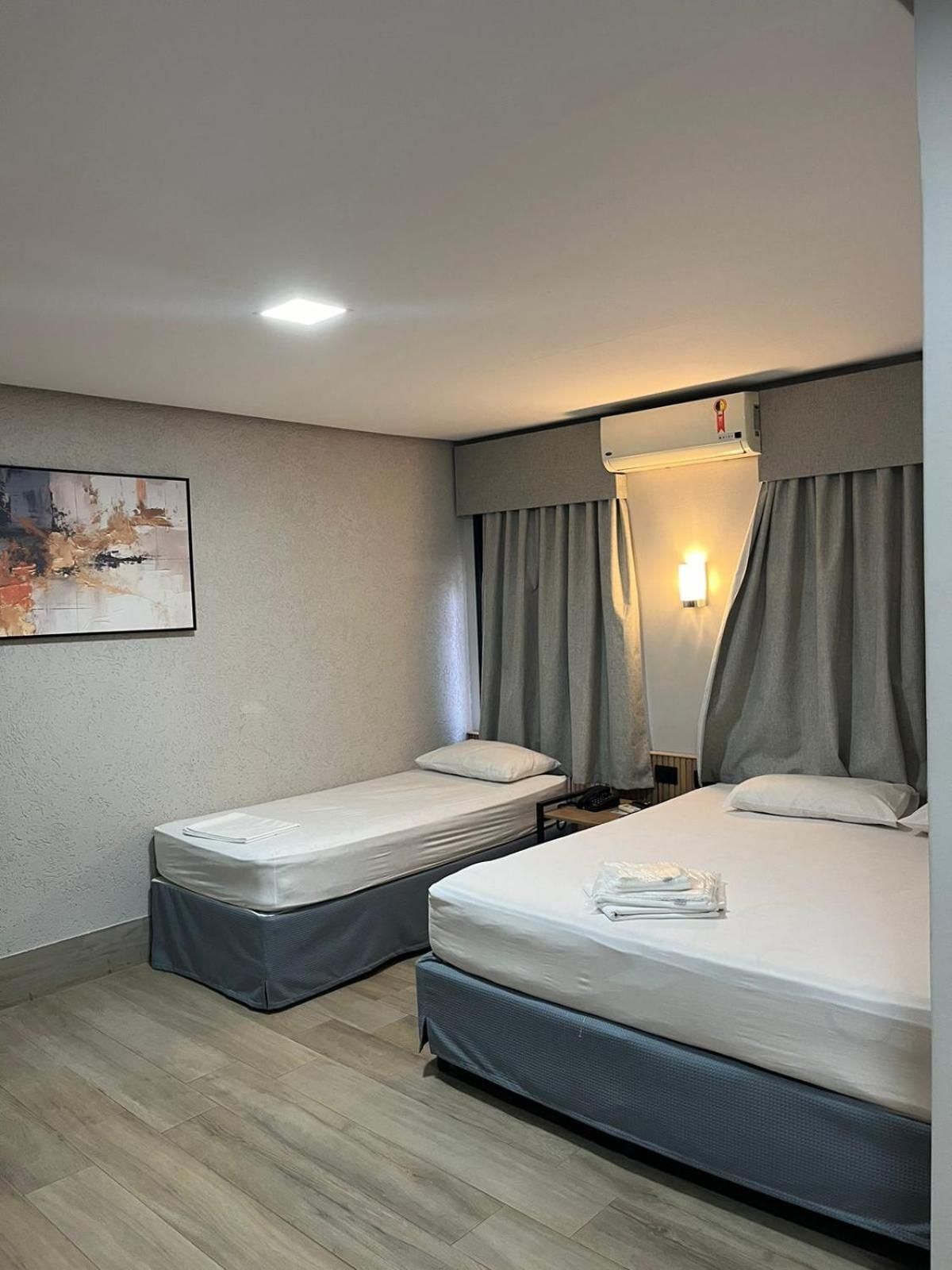 Quarto de hotel com duas camas de solteiro, mesa de cabeceira com abajur, ar-condicionado fixado na parede e um quadro na parede.