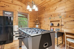 A wooden-floored room with a foosball table, a black refrigerator, and a window with blinds. - Seven Feathers A Modern Barn Retreat/ Pet & Family Friendly (Blue Ridge)