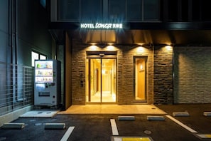 Entrada de hotel con una máquina expendedora, fachada de piedra y un cartel sobre las puertas.