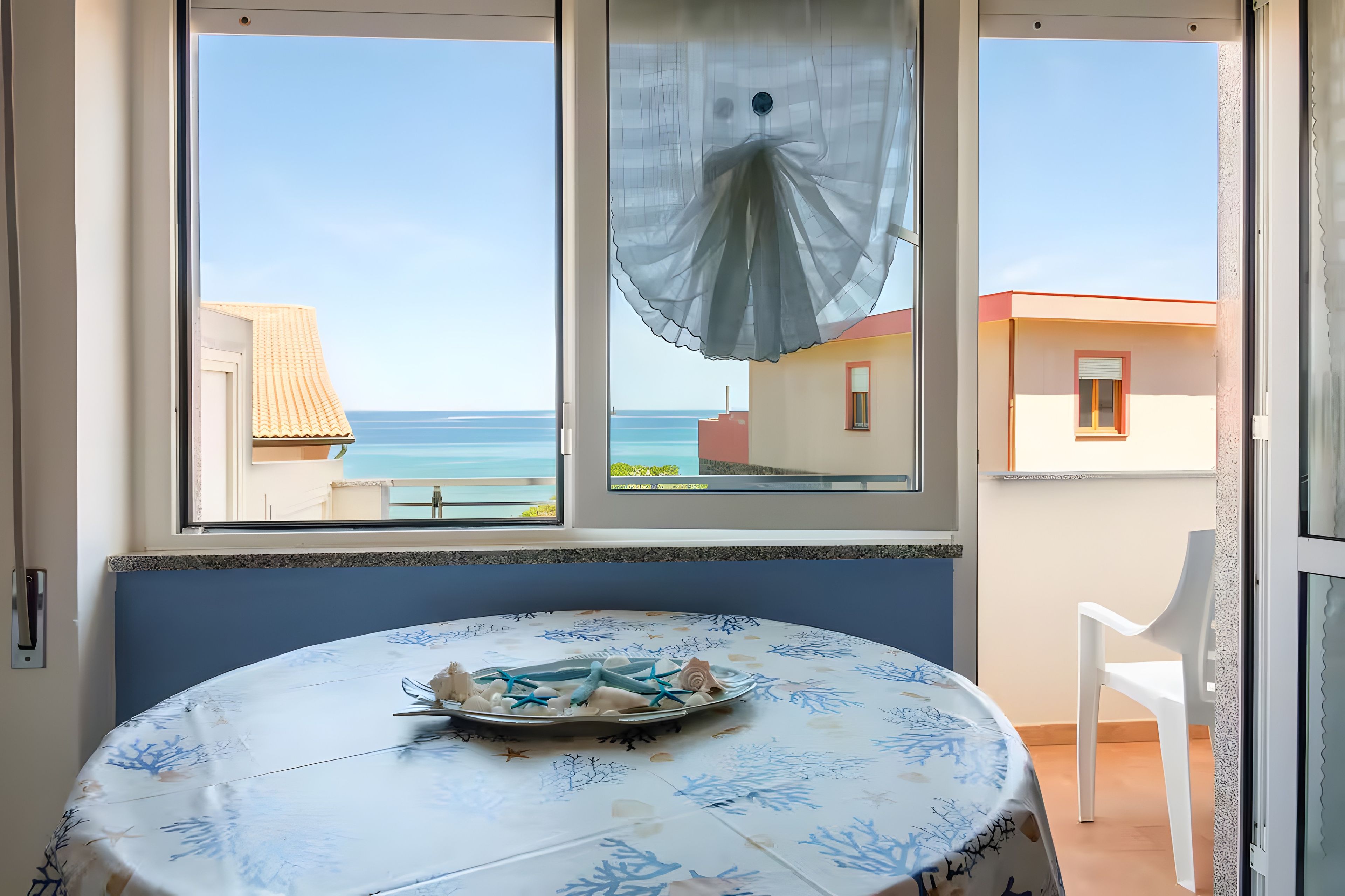 Et værelse med et bord dækket til to, havudsigt gennem et vindue og en balkon med en stol.