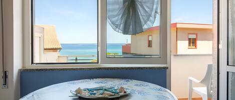 A room with a table set for two, a view of the sea through a window, and a balcony with a chair.