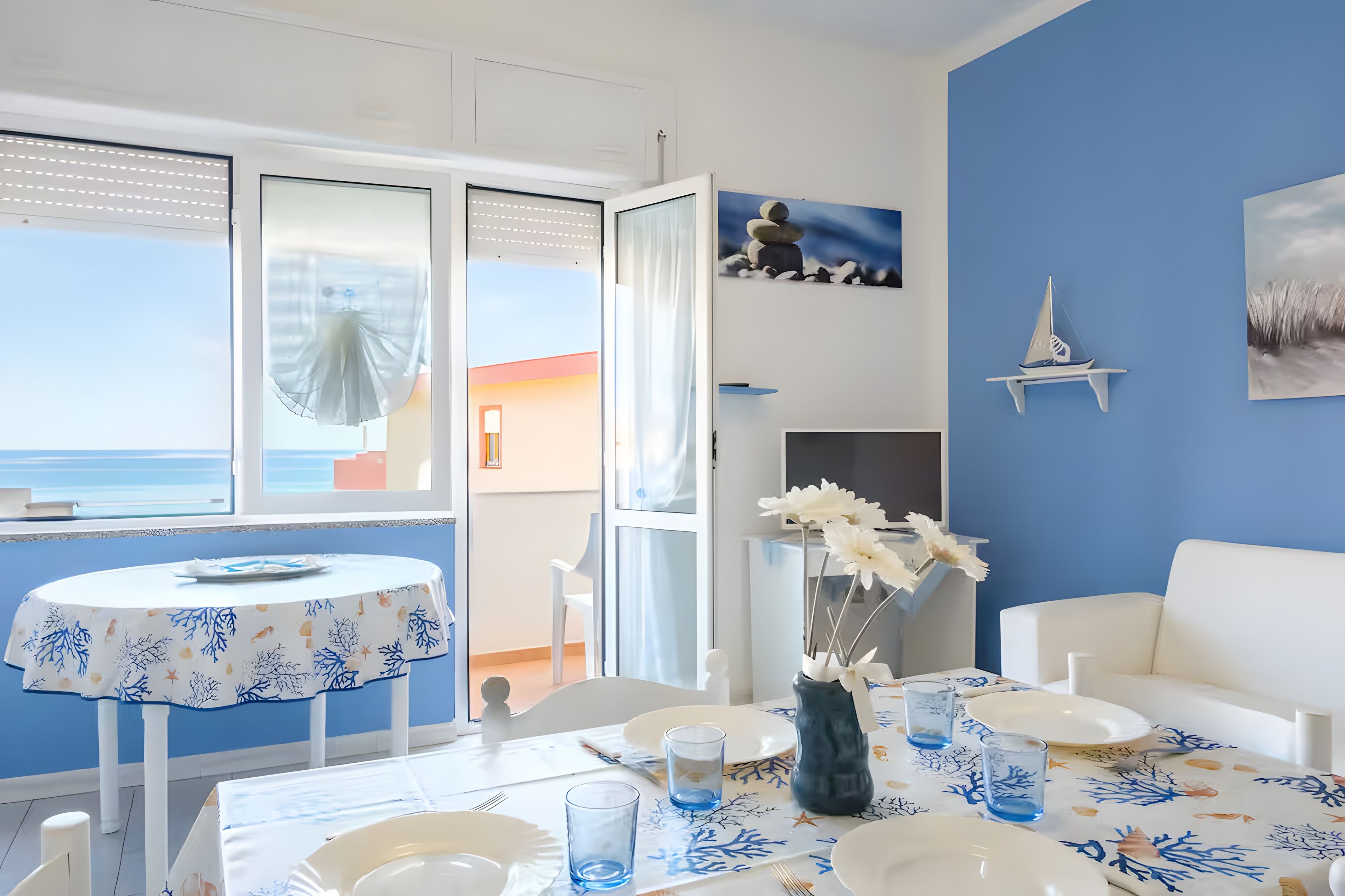 Ein Essbereich mit einem Tisch für zwei Personen, Meerblick durch ein Fenster und eine blaue Wand mit einem Segelbootgemälde.