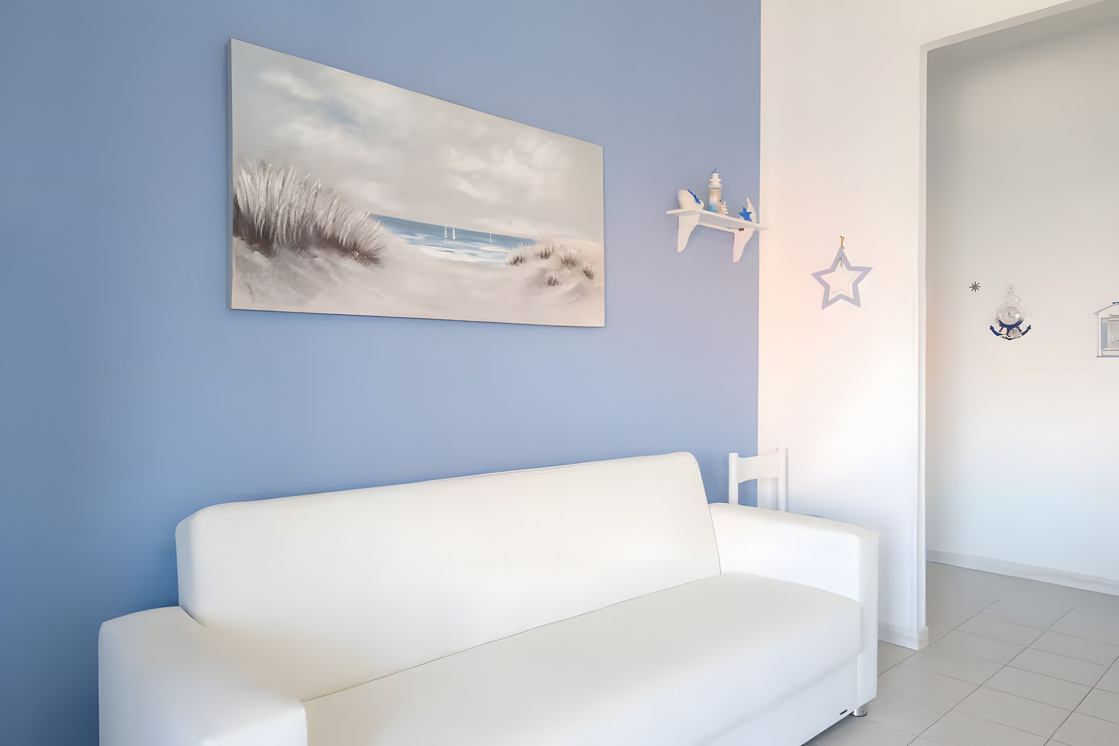 A white sofa against a blue wall with a beach scene painting, a star decoration, and a small shelf with items.