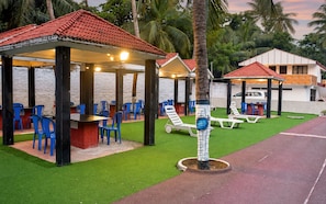 A covered outdoor seating area with blue chairs and tables, surrounded by palm trees and a white building.