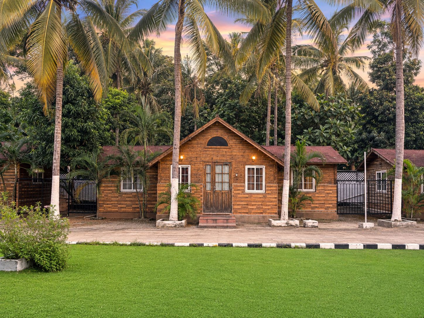 A wooden bungalow with a central door, surrounded by lush greenery and palm trees.