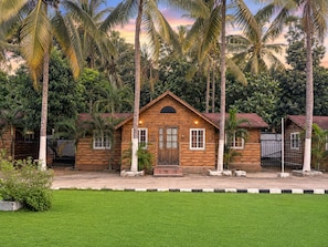A wooden bungalow with a central door, surrounded by lush greenery and palm trees.