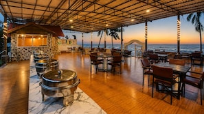 Restaurante com terraço virado para o mar, com chão de madeira, panelas de metal e uma parede de pedra com um mural.
