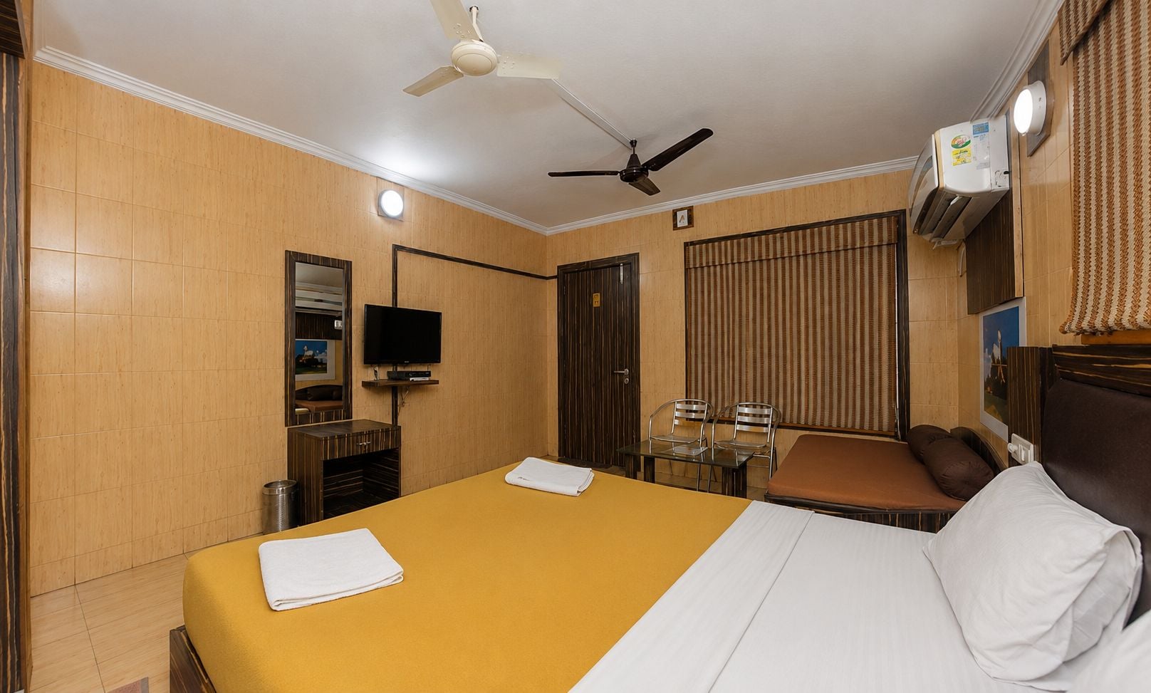 A hotel room with a bed, a television, a desk with chairs, and a ceiling fan.