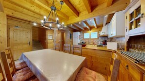A wooden interior with a dining table, chairs, a chandelier, and a kitchen area.