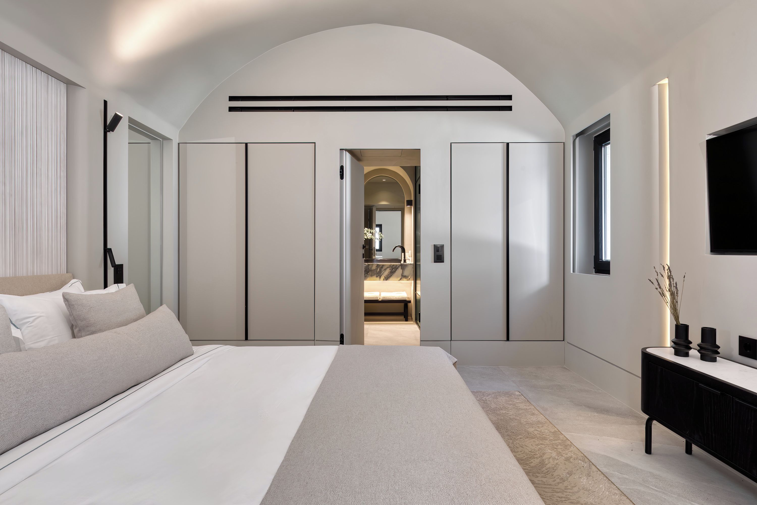 A modern bedroom with a large bed, a walk-in closet, and a bathroom visible through an archway.