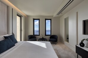 Une chambre moderne avec un grand lit, deux fenêtres donnant sur l’océan, une petite table et une chaise.