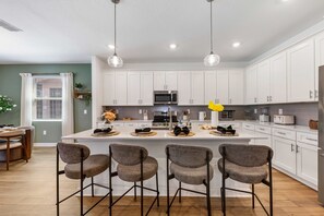 A modern kitchen with white cabinets, a central island with bar stools, and pendant lighting. - Brand New Family 6BR Pool Spa Game Lounge 7875 (Kissimmee)