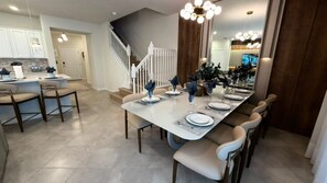A modern dining area with a long table set for six, a kitchen in the background, and a staircase leading to another level. - Brand New Modern 5BR With Pool Game Room 16212 (Clermont)