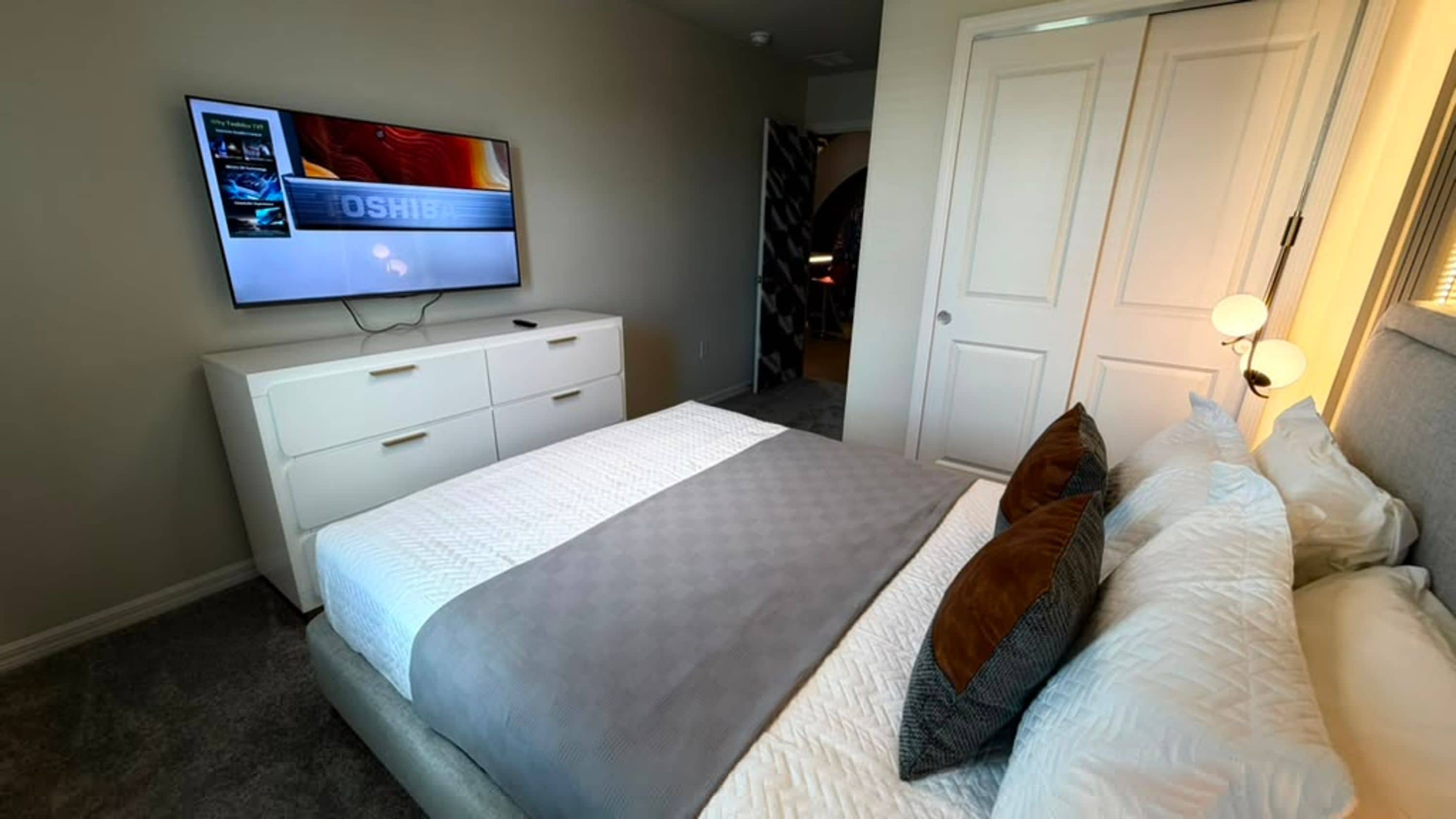 A hotel room with a bed, a television mounted on the wall, a white dresser, and a door leading to another room.