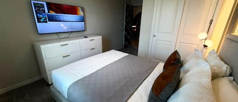 A hotel room with a bed, a television mounted on the wall, a white dresser, and a door leading to another room.