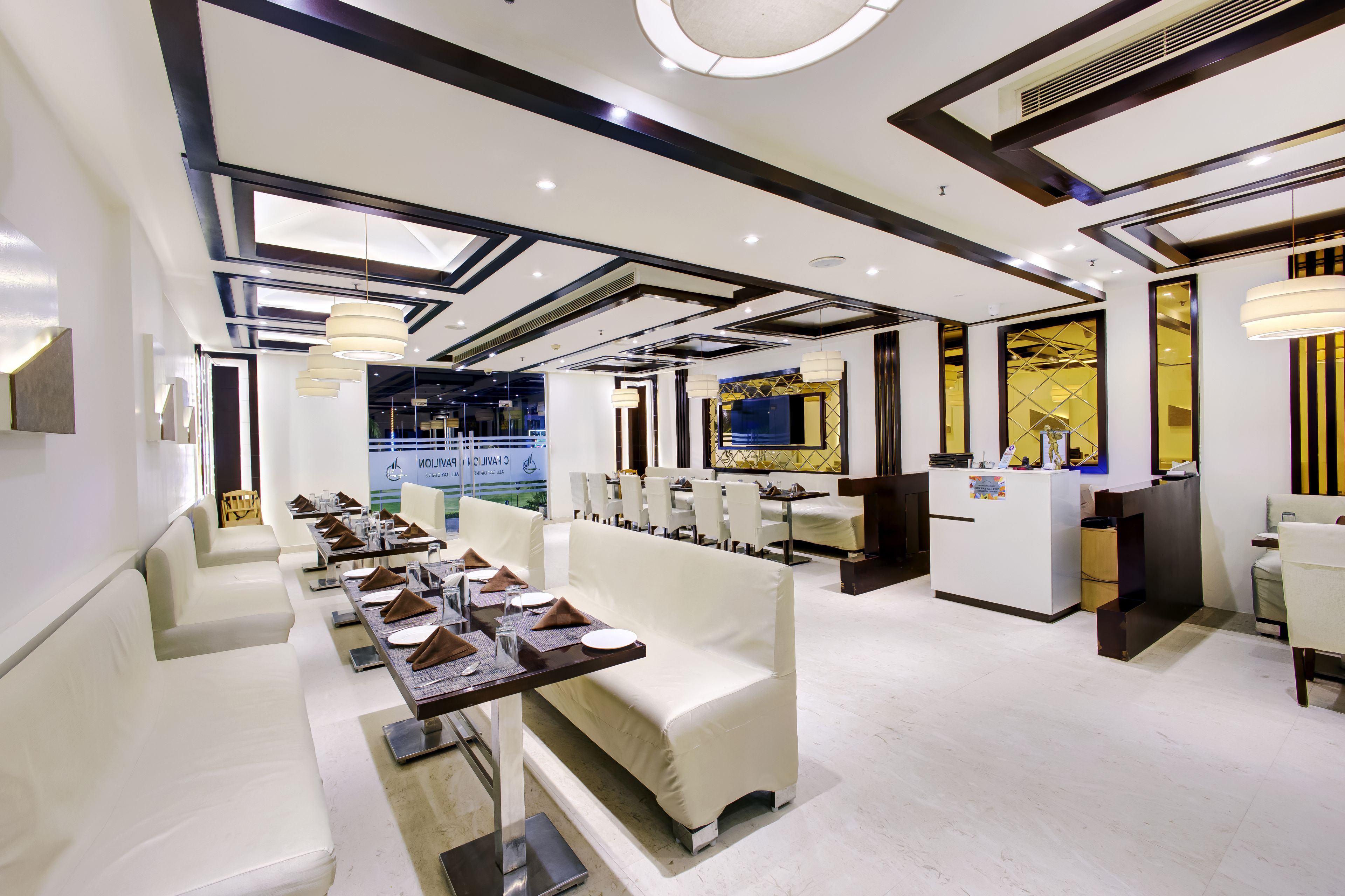 Interior moderno de restaurante com mobiliário branco, iluminação embutida e área de jantar central.