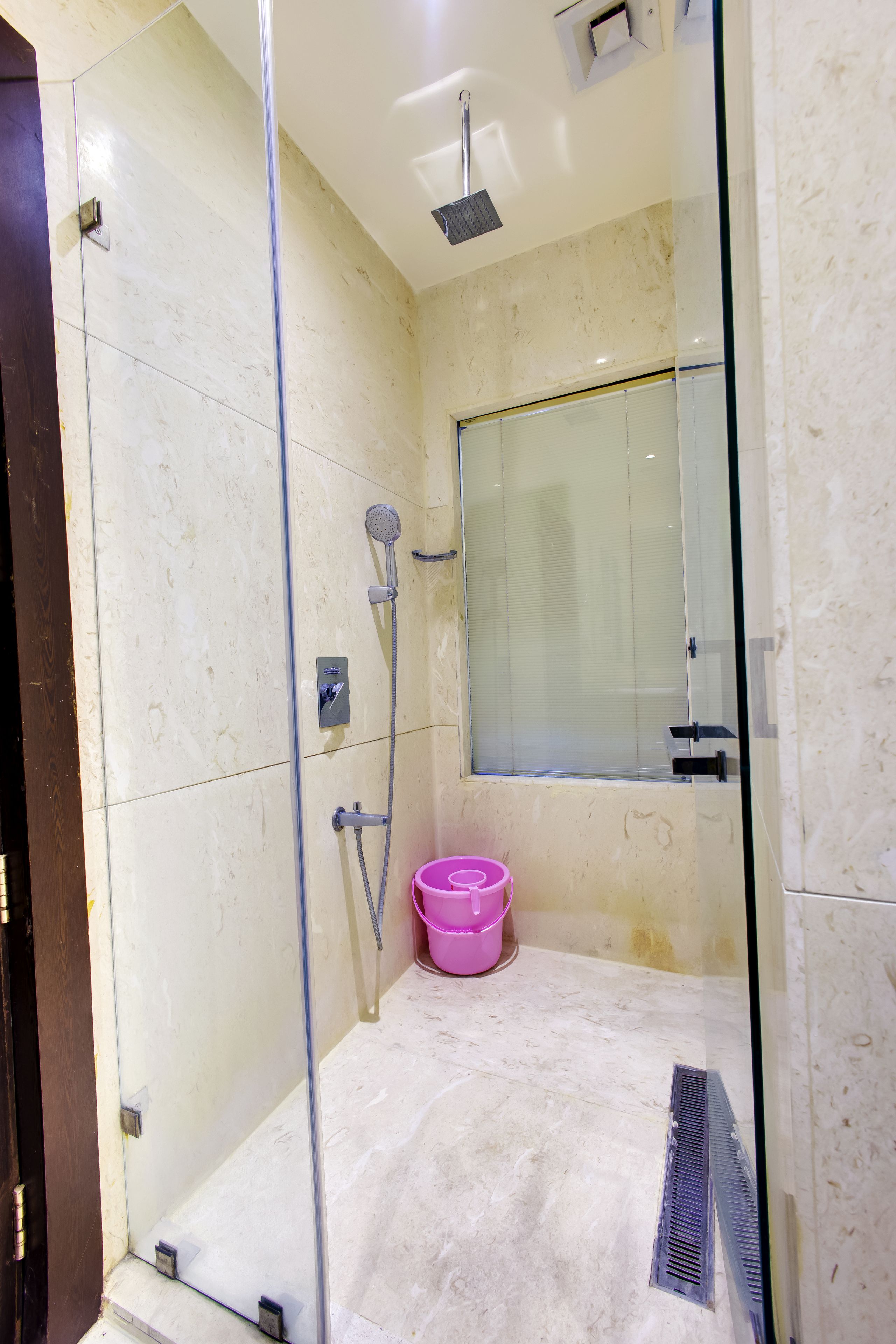 A glass-enclosed shower with a handheld showerhead, a pink bucket, and a ventilation grate.