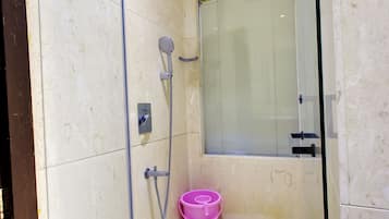 A glass-enclosed shower with a handheld showerhead, a pink bucket, and a ventilation grate.