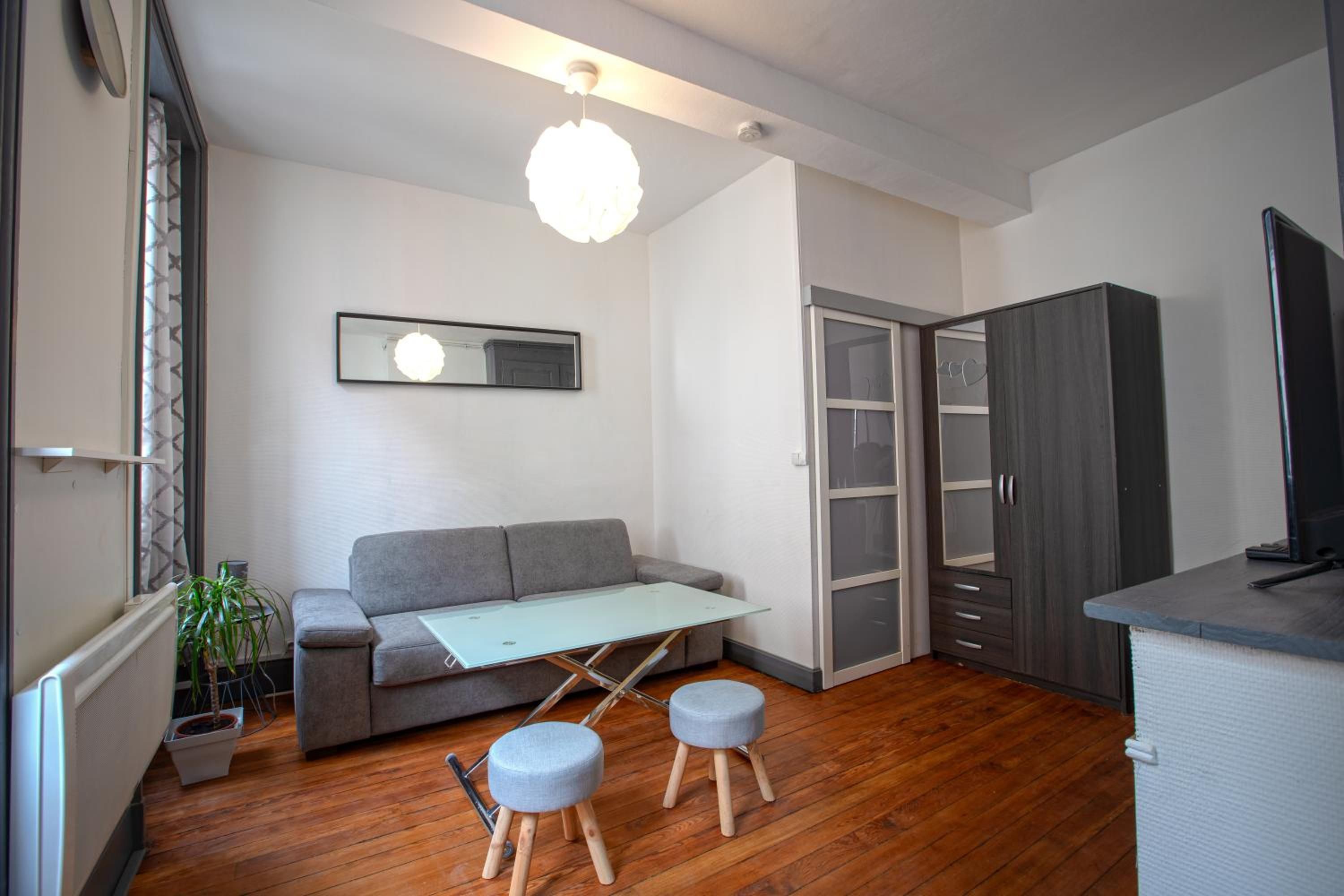 Un espace de vie moderne comprenant un canapé gris, une petite table, deux tabourets, un miroir et une armoire.