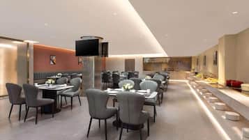 A modern restaurant with multiple dining tables and chairs, a flat-screen TV mounted on the wall, and a buffet area with various dishes.