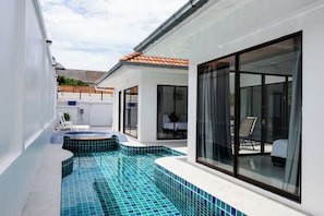 Una piscina exterior moderna con suelo de baldosas, zona de estar cubierta y una habitación con cama, completamente cerrada con vidrio.
