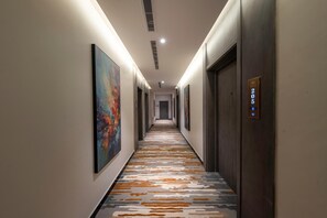 A hotel hallway with a patterned carpet, framed artwork on the walls, and a door with a number plaque. - Gardn Millennium Al-salamah Hotel Jeddah (Jeddah)