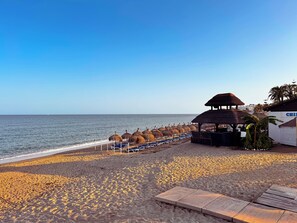 Une plage avec des chaises longues et des parasols en chaume, une terrasse en bois et un bâtiment sur lequel on peut voir une enseigne.