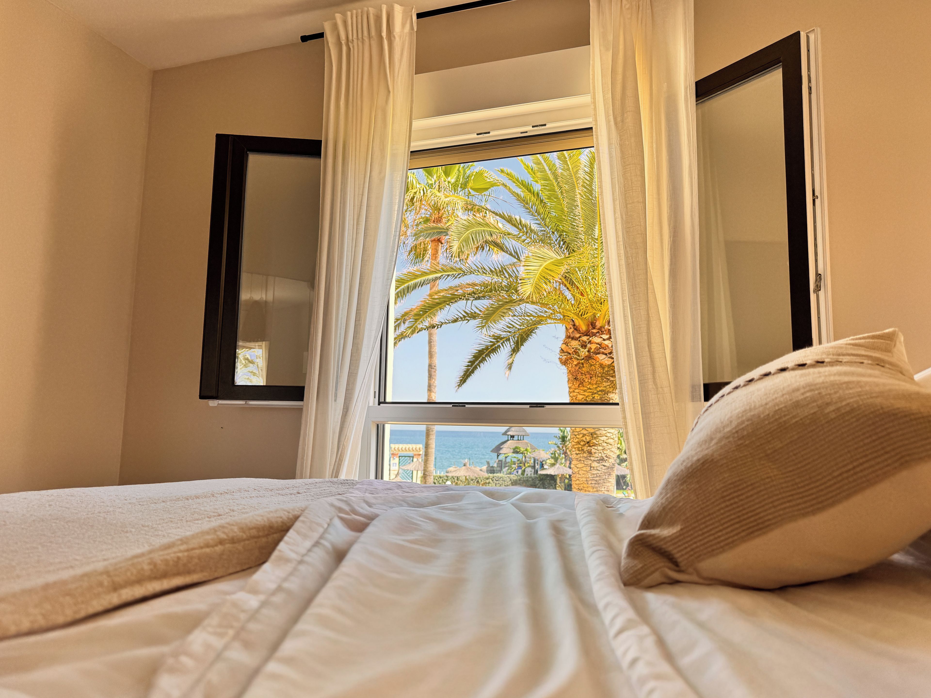 A bed with white bedding and a pillow, a window with white curtains, and a view of palm trees and a building.
