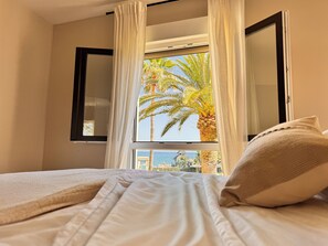 A bed with white bedding and a pillow, a window with white curtains, and a view of palm trees and a building. - Beachfront • Sea Views • The Mimosas • La Cala de Mijas (La Cala de Mijas)