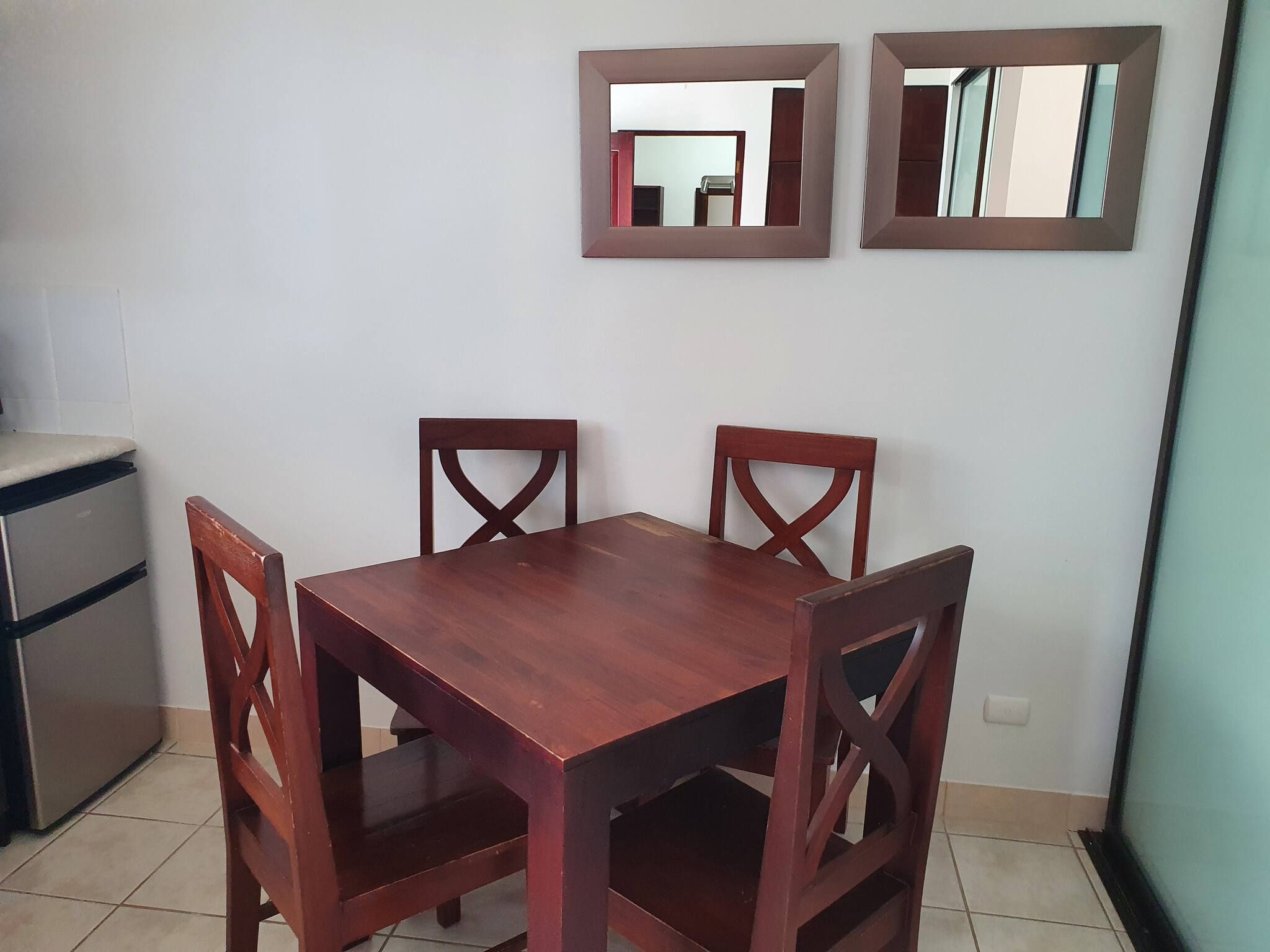 Un comedor con mesa de madera y cuatro sillas, un refrigerador y dos espejos fijados a la pared.