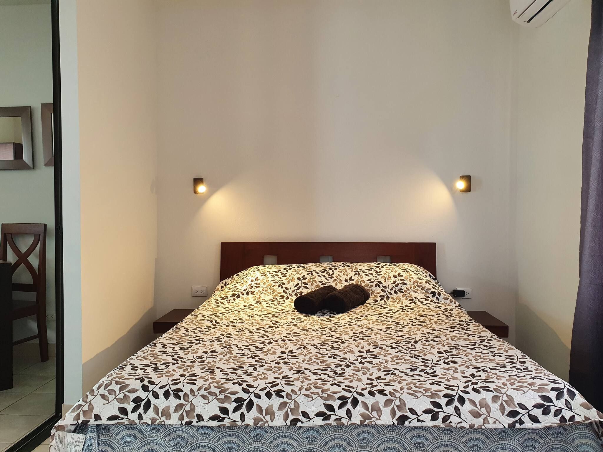 Una cama individual con una colcha estampada, dos lámparas fijadas a la pared, un cabecero de madera y un enchufe en la pared.