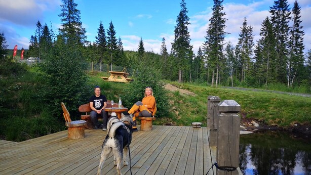 En træterrasse med to personer siddende ved et bord, en hund i snor og en lille sø i baggrunden.