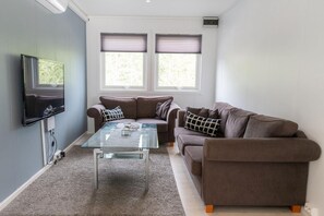 A living room with a flat-screen TV mounted on the wall, two sofas, a glass coffee table, and a window with blinds. - Blafjell Hundesenter Fjellhotell (Vefsn)