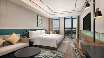 A modern hotel room with a bed, sofa, TV, and a view of the outdoors.