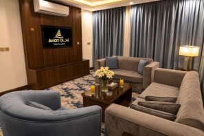 Habitación de hotel con una televisión de pantalla plana montada en un panel de madera, un conjunto de sofás, una mesa de centro con un florero y una lámpara.