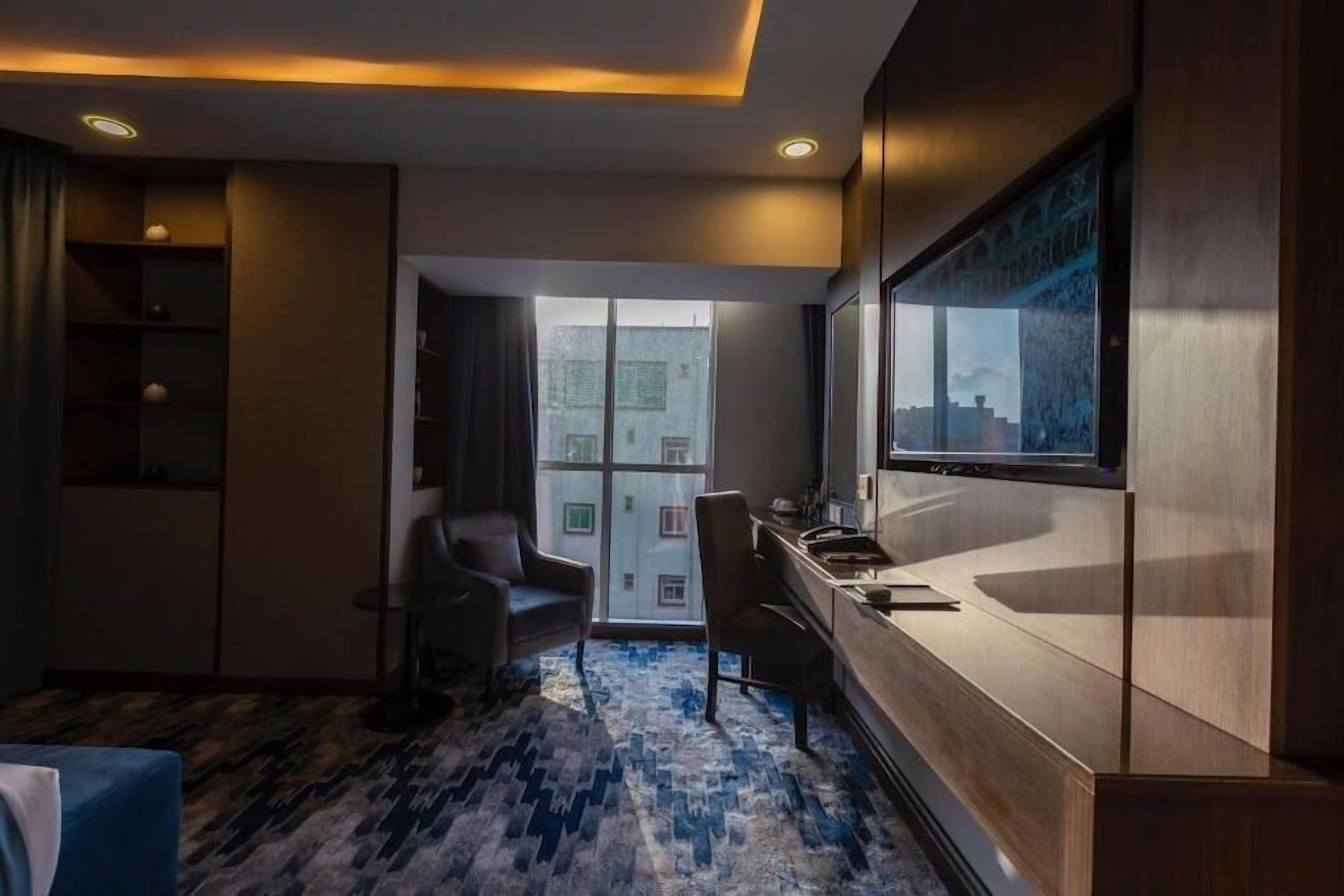 A hotel room with a large window, a desk, a chair, and a patterned carpet.